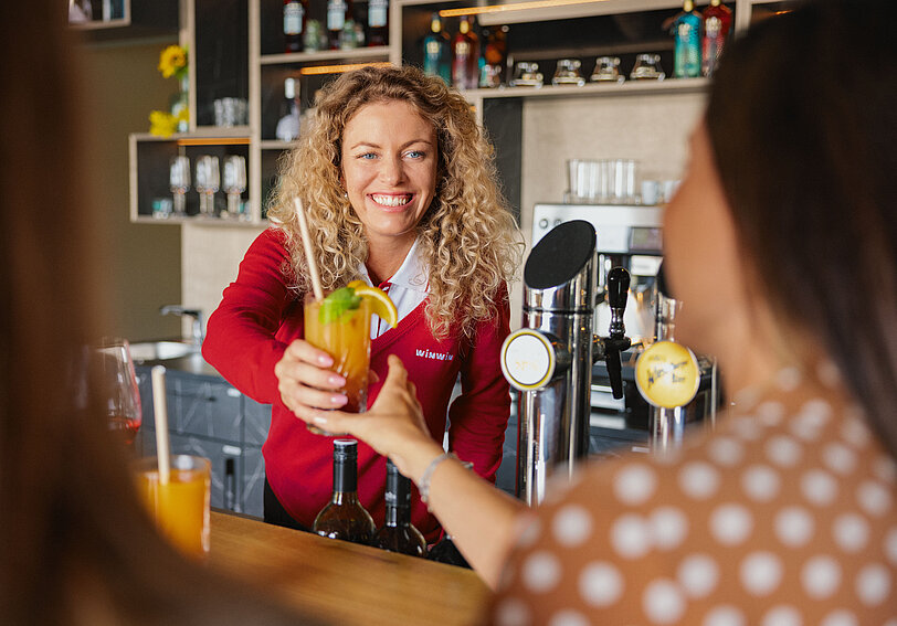 Blond gelockte WINWIN Mitarbeiterin serviert Cocktails an zwei Damen an der Bar im WINWIN Sankt Poelten.