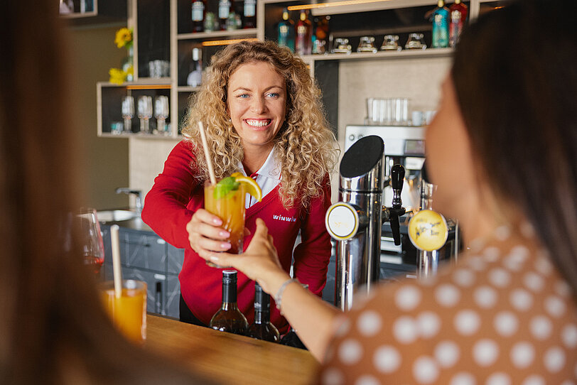 Blond gelockte WINWIN Mitarbeiterin serviert Cocktails an zwei Damen an der Bar im WINWIN Sankt Poelten.