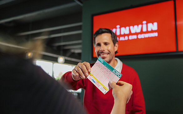 Kundin ueberreicht WINWIN Mitarbeiter einen ausgefuellten Lotto-Schein.