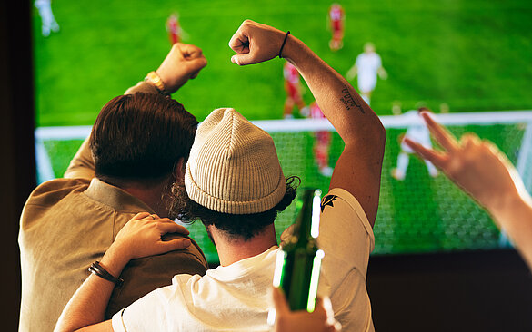 Gaeste jubeln vor TV bei einem Fußballmatch. Mann mit Muetze freut sich mit Freund.