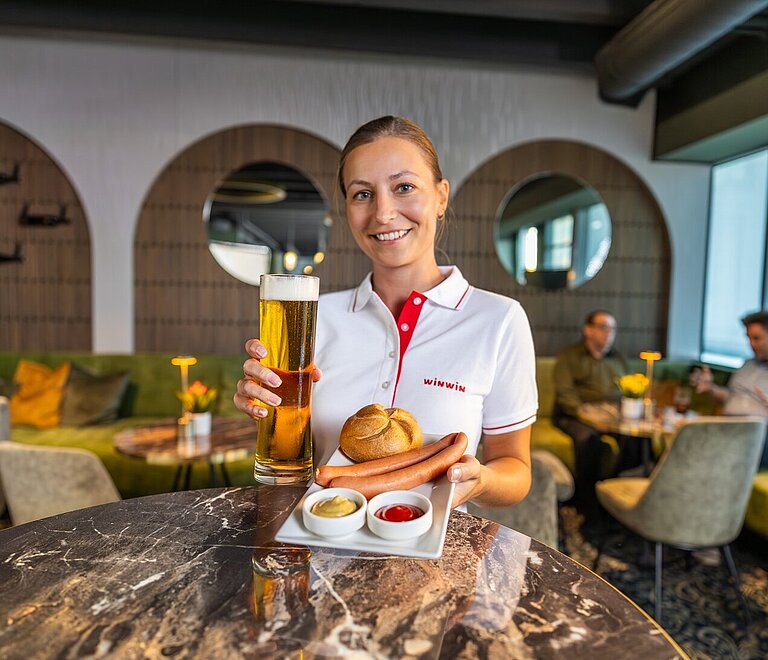Eine junge Frau in WINWIN-Dienstkleidung hält ein Paar Würstel und ein Glas Bier.