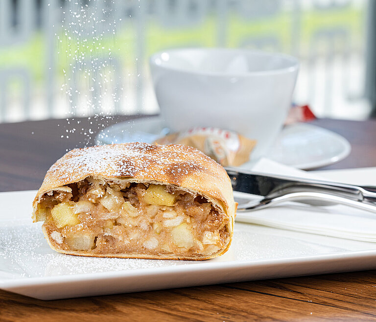 Ein großes Stueck Apfelstrudel wird mit Staubzucker bestreut. Tasse Kaffee im Hintergrund.