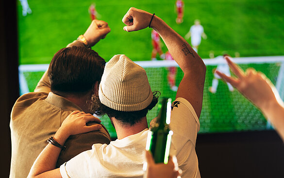 Gaeste jubeln vor TV bei einem Fußballmatch. Mann mit Muetze freut sich mit Freund. 