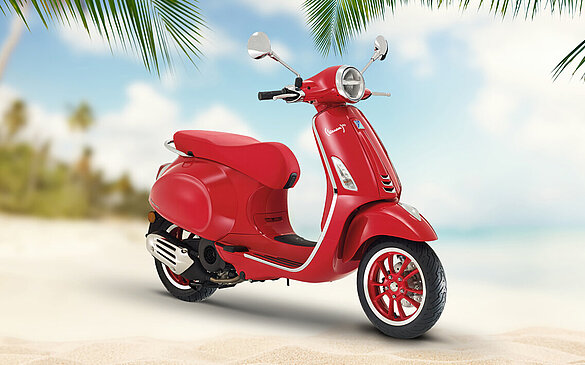 Eine rote Vespa steht an einem sonnigen Sandstrand.