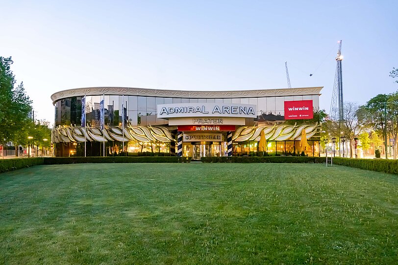WINWIN Prater Arena Eingang mit beleuchteten Glasfronten und Fahrgeschaeft im Hintergrund. 