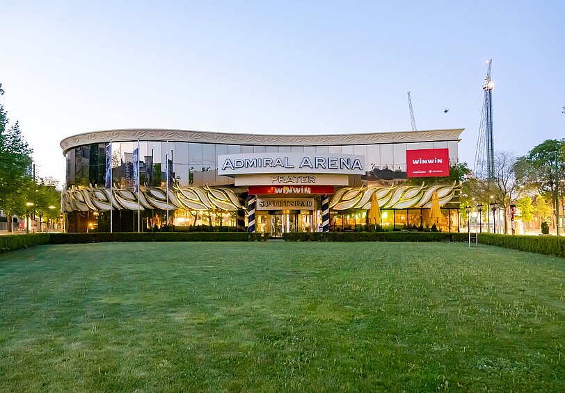 WINWIN Prater Arena Eingang mit beleuchteten Glasfronten und Fahrgeschaeft im Hintergrund. 