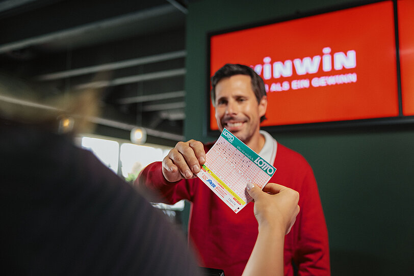Kundin ueberreicht WINWIN Mitarbeiter einen ausgefuellten Lotto-Schein.