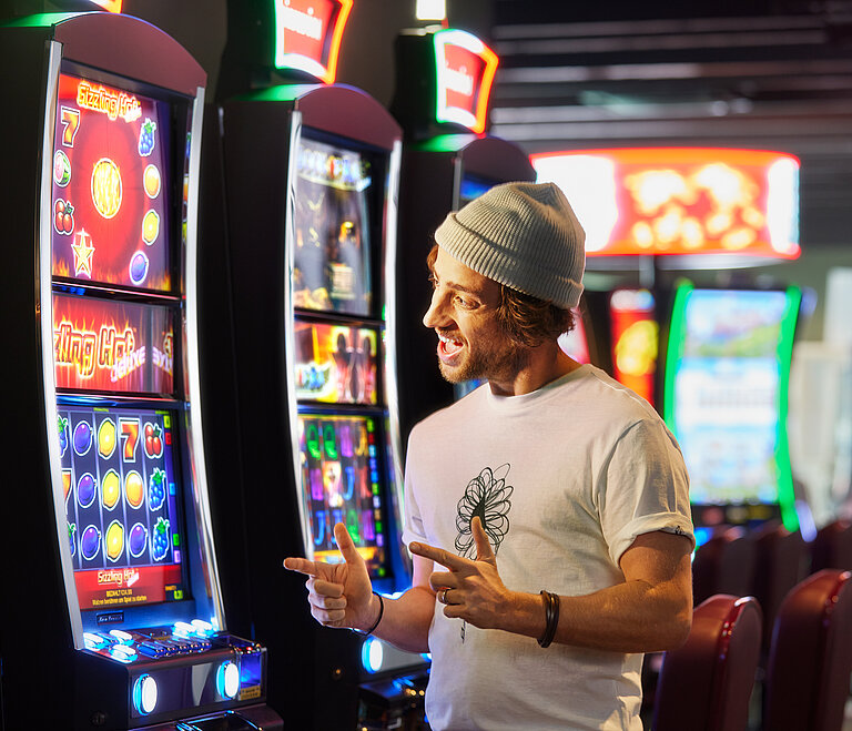 Junger Mann mit weißer Muetze freut sich im Spielbereich und zeigt auf einen VLT.