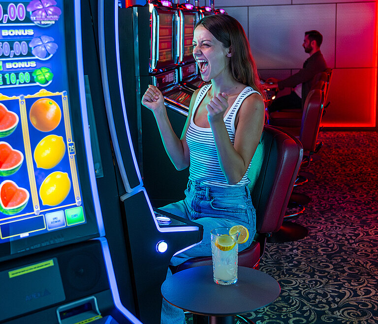 Eine junge Frau in legerer Kleidung sitzt vor einem Spielautomaten und jubelt.