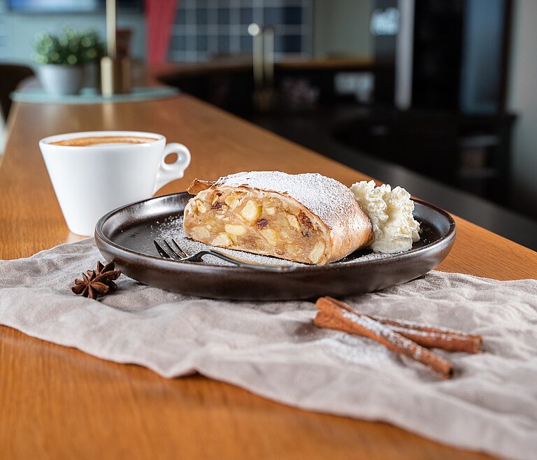 Apfelstrudel auf einem schwarzen Teller, daneben eine Tasse Kaffee