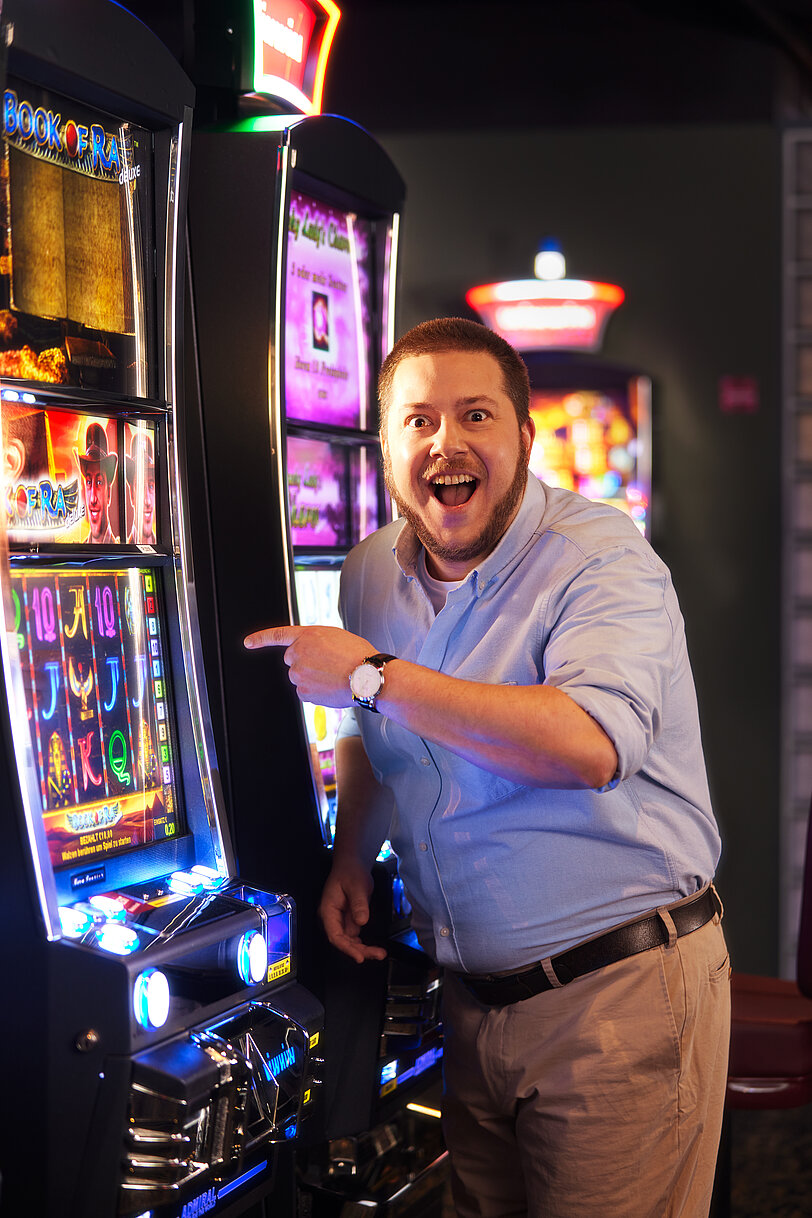 Mann mit blauem Hemd steht im Spielbereich und zeigt freudig auf einen VLT.