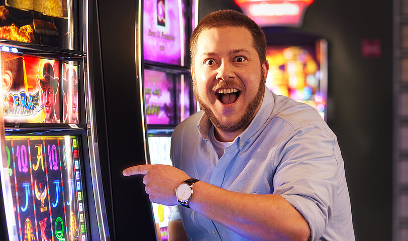 Mann mit blauem Hemd steht im Spielbereich und zeigt freudig auf einen VLT.