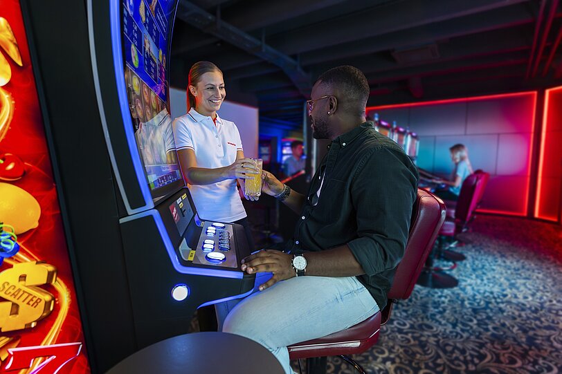 WINWIN Mitarbeiterin überreicht einem Gast sein Getränk während er am Spielautomat sitzt