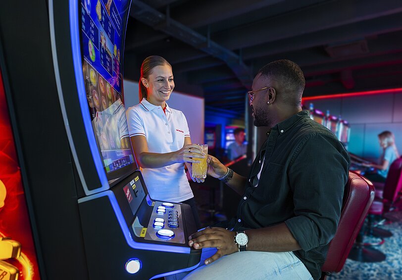 WINWIN Mitarbeiterin überreicht einem Gast sein Getränk während er am Spielautomat sitzt