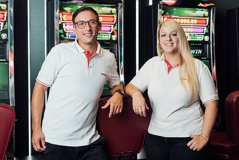 Ein männlicher WINWIN Mitarbeiter und eine weibliche WINWIN Mitarbeiterin stehen nebeneinander vor Spielautomaten und lächeln in die Kamera