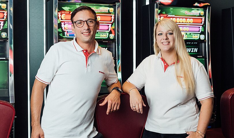 Ein männlicher WINWIN Mitarbeiter und eine weibliche WINWIN Mitarbeiterin stehen nebeneinander vor Spielautomaten und lächeln in die Kamera