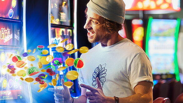 Junger Mann mit Haube zeigt auf Spielautomat mit fliegenden Slot Game-Fruechten.