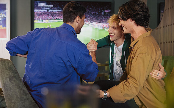 Drei Freunde schauen gemeinsam ein Fußballspiel im WINWIN und freuen sich miteinander.